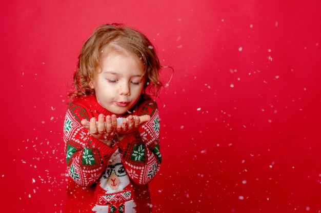 Niña en un suéter con renos soplando nieve sobre un fondo rojo Navidad año nuevo