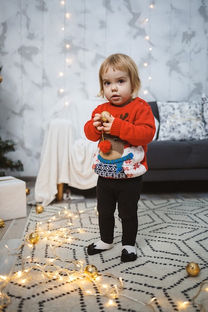 Una niña con un suéter rojo cálido se encuentra cerca de un árbol de Navidad con juguetes y regalos. Infancia feliz. Ambiente de vacaciones de año nuevo