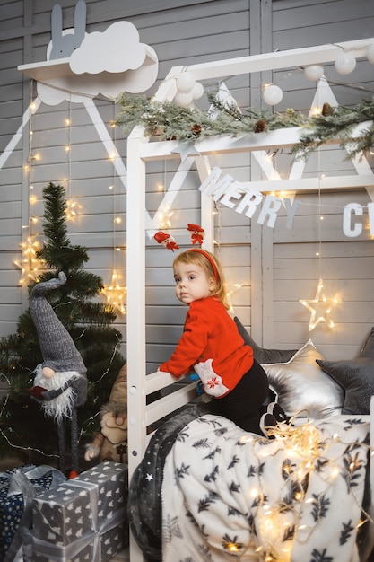 Una niña con un suéter rojo cálido se sienta en una cama cerca de un árbol de Navidad con juguetes y regalos. Infancia feliz. Ambiente de vacaciones de año nuevo