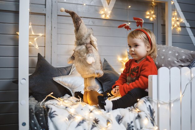 Una niña con un suéter rojo cálido se sienta en una cama cerca de un árbol de Navidad con juguetes y regalos. Infancia feliz. Ambiente de vacaciones de año nuevo
