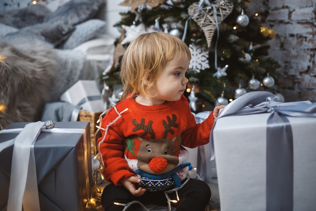 Una niña con un suéter rojo cálido se sienta debajo de un árbol de Navidad con juguetes y regalos. Infancia feliz. Ambiente de vacaciones de año nuevo