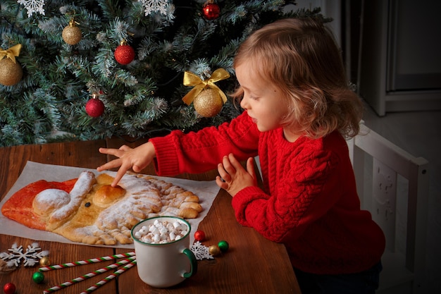 Una niña con un suéter rojo toca un pastel de Navidad en forma de Santa en una mesa con adornos