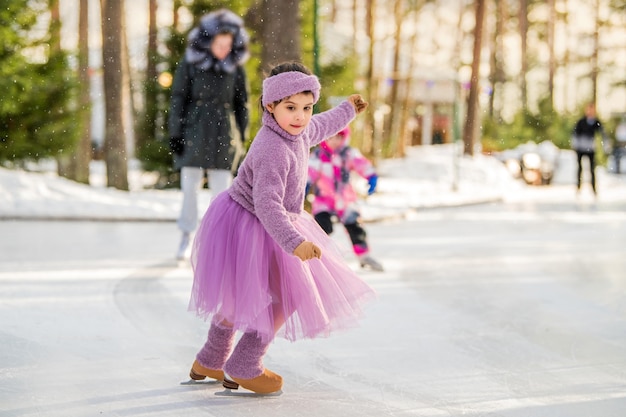 Niña con un suéter rosa y una falda completa paseos en un día soleado de invierno en una pista de hielo al aire libre en el parque