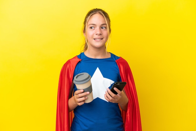 Niña superhéroe aislada sobre fondo amarillo sosteniendo café para llevar y un móvil mientras piensa en algo