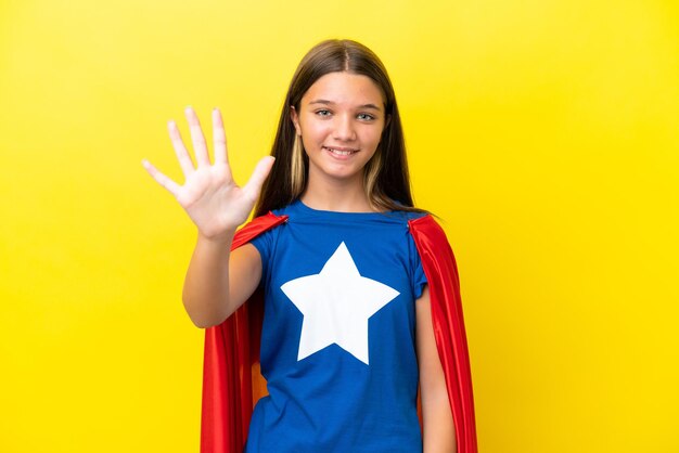 Niña superhéroe caucásica aislada de fondo amarillo contando cinco con los dedos