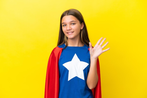 Niña superhéroe caucásica aislada de fondo amarillo saludando con la mano con expresión feliz