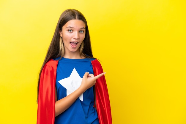 Niña superhéroe caucásica aislada de fondo amarillo sorprendida y apuntando hacia el lado