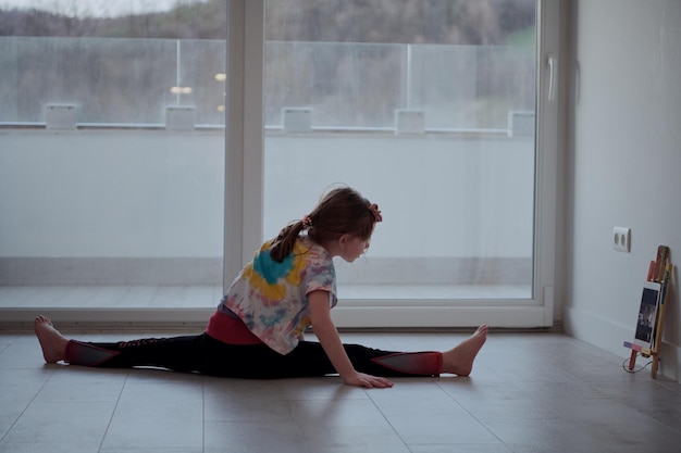 La niña tiene entrenamiento en línea a través de una tableta y hace ejercicios de danza de ballet moderno en casa. Clase conceptual de educación en línea en la escuela de ballet moderna. Distancia social durante la cuarentena, autoaislamiento