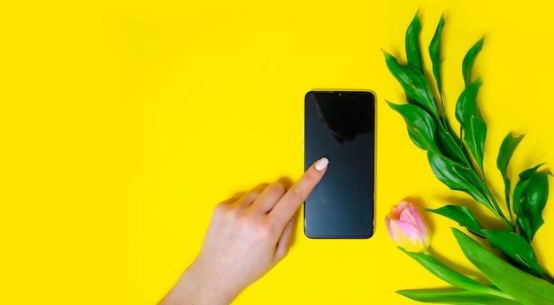 La niña tiene el teléfono en la mano. Hojeando las páginas. Señalando el teléfono. Flores Fondo amarillo brillante. Lugar para una inscripción. Endecha plana.