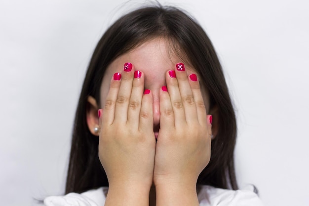 Niña tímida se cubre la cara con las manos mostrando uñas de esmalte rosa sobre fondo blanco.