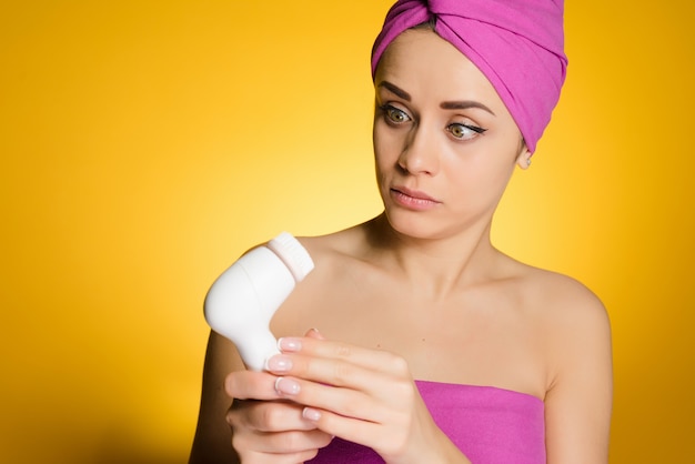 Una niña con una toalla rosa en la cabeza sostiene un cepillo eléctrico para limpiar su cara y parece sorprendida.