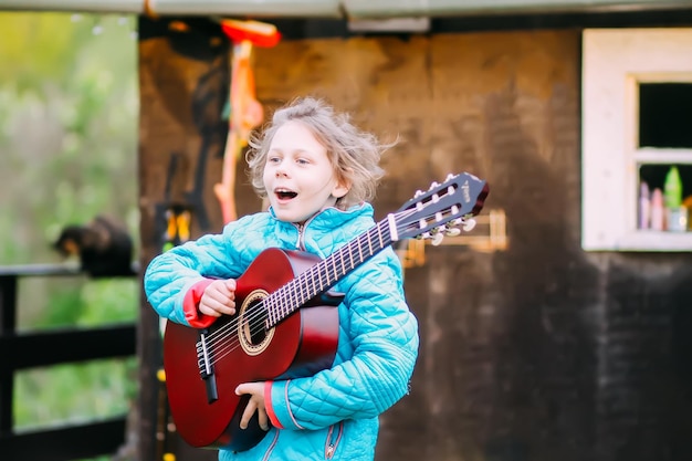 Niña tocando la guitarra y cantando al aire libre en el prado verde en primavera Niño feliz con instrumento musical en el patio rural