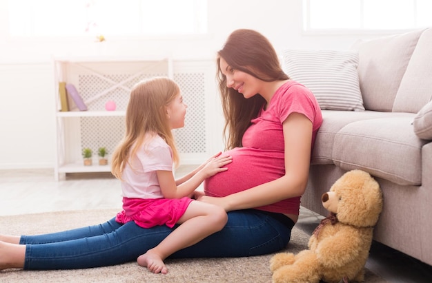 Niña tocando su vientre de mamá embarazada. Esperando a la madre sentada con su hija en el piso en casa, divirtiéndose y sintiendo los empujones del bebé, copie el espacio