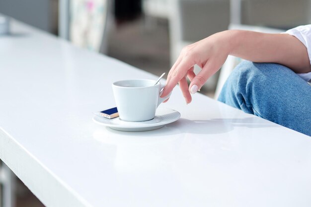 La niña toma una taza de café en la mano de una mesa blanca en un café.