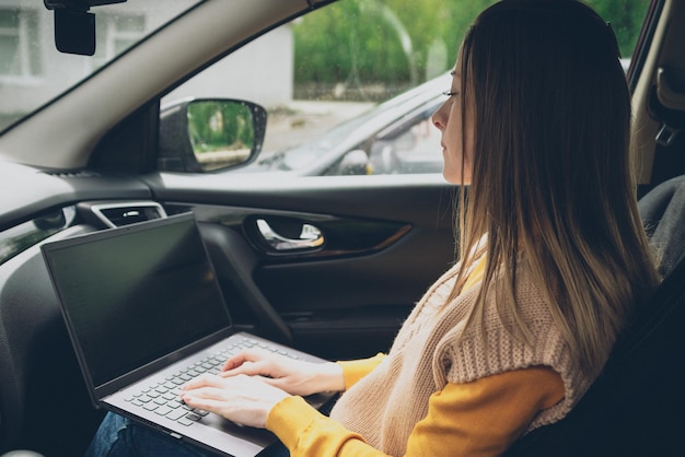 Una niña trabaja en una computadora portátil mientras está sentada en un automóvil, una trabajadora independiente o bloguera en el trabajo, trabajo remoto, tecnologías modernas