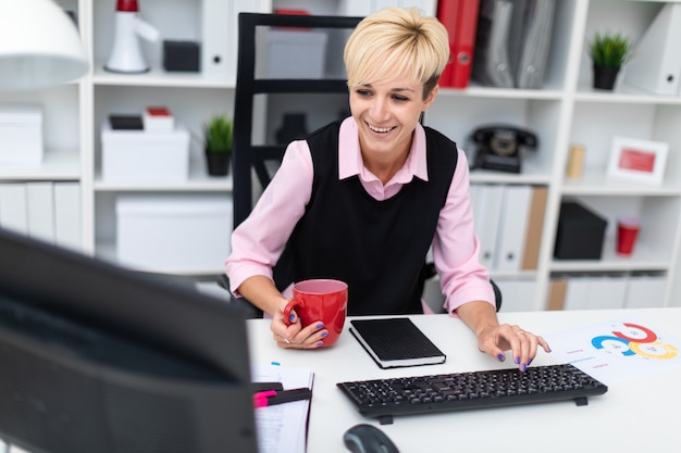 La niña trabaja en la oficina en la computadora y sostiene una taza.