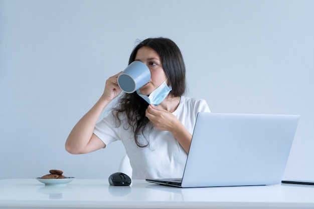 La niña trabaja desde su casa durante la cuarentena. Pandemia de coronavirus. Una niña con una máscara quirúrgica trabaja en una computadora portátil y toma café. Trabajo distante