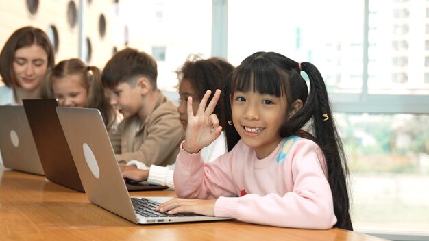 Niña trabajando en la computadora portátil y mirando o haciendo ok o ok en la cámara Erudición