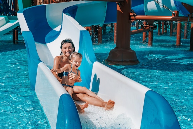 Niña en traje de baño baja los toboganes azules a la piscina, mamá e hija juegan y nadan en la piscina al aire libre