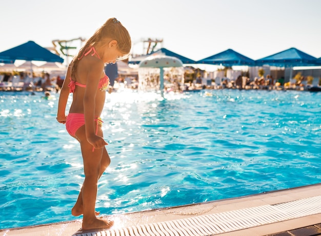 Una niña en un traje de baño de color rosa intenso está junto a la piscina con agua clara y transparente y se prueba el pie