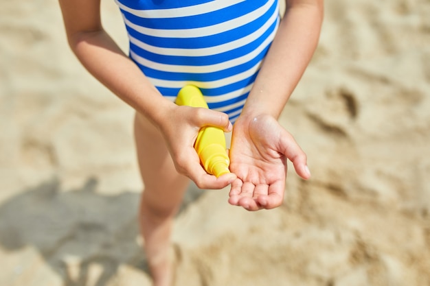 La niña en traje de baño sostiene en la mano una botella amarilla de protector solar de pie en la playa, protección solar para la piel. Vacaciones de verano.