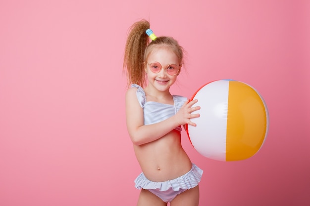 Niña en traje de baño sostiene una pelota de vacaciones sobre un fondo rosa