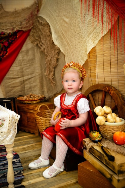 Una niña con un traje nacional rojo cerca de una mesa con un samovar y volantes Día de panqueques