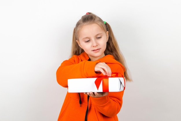 Una niña con traje naranja tiene un regalo con una cinta roja en un fondo blanco