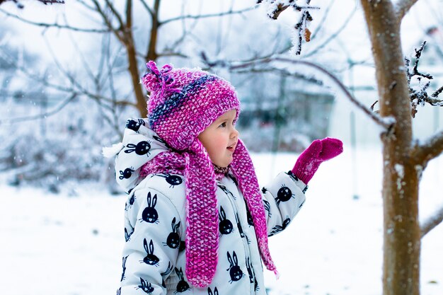 Niña en traje de nieve blanco y sombrero rosa, botas y guantes en el parque de invierno de nieve.