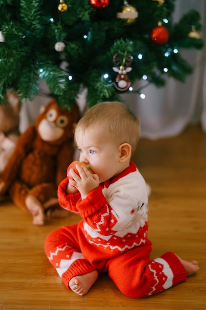 Una niña con un traje de punto rojo y blanco muerde una manzana frente a un árbol de Navidad.