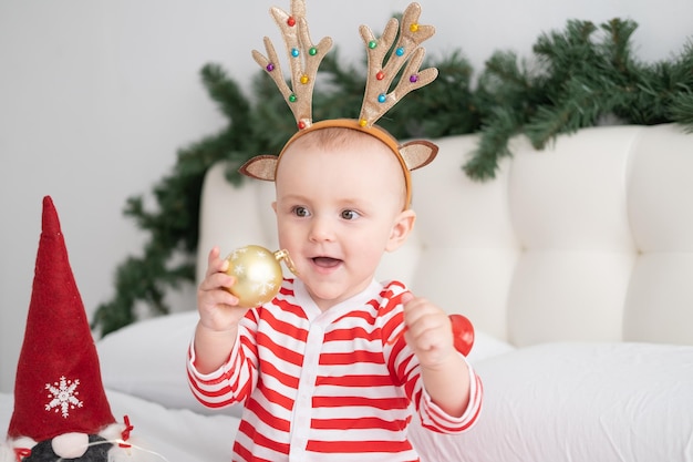 Niña en traje de rayas con cuernos de ciervo en la cabeza jugando con bolas de navidad en la cama decorada