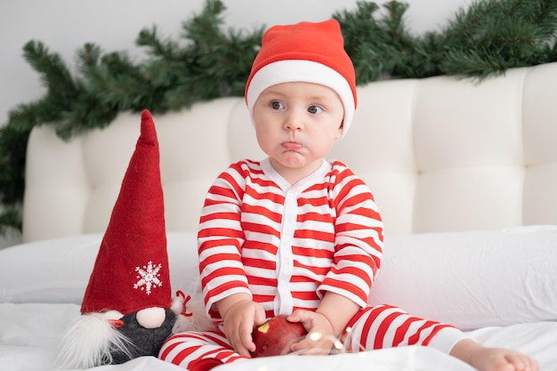 Niña en traje de rayas y gorro de Papá Noel comiendo manzana roja en la cama decorada de Navidad