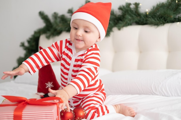 Niña en traje de rayas y gorro de Papá Noel jugando con caja de regalo en dormitorio decorado de Navidad
