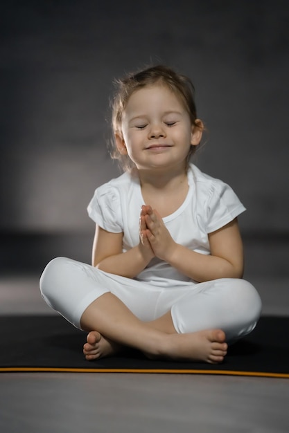 Niña de tres años meditando en una pose de loto sobre un fondo gris en una habitación oscura