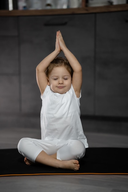 Niña de tres años meditando en una pose de loto sobre un fondo gris en una habitación oscura