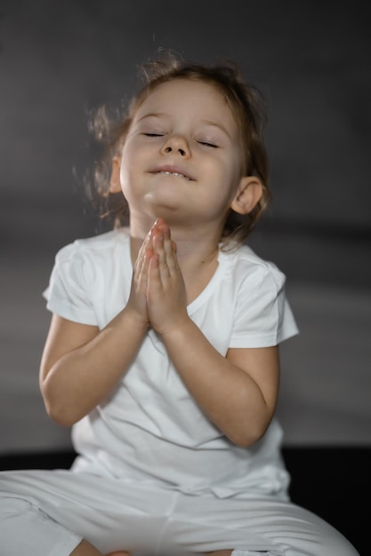 Foto niña de tres años meditando en una pose de loto sobre un fondo gris en una habitación oscura