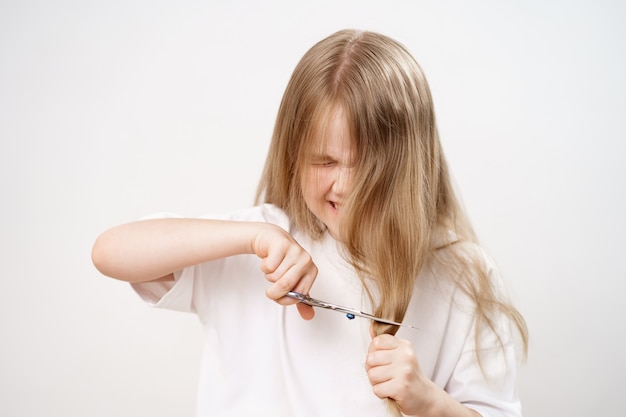 La niña triste corta el pelo largo con unas tijeras sobre un fondo blanco. Corte de pelo de moda para el bebé. Peluquero. travesuras de los niños. Corte de pelo