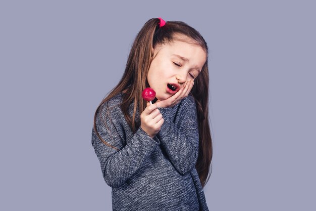 Niña triste con una paleta roja en sus manos, cuyos dientes duelen. El concepto de desarrollo de caries debido al abuso de dulces. aislado en superficie gris