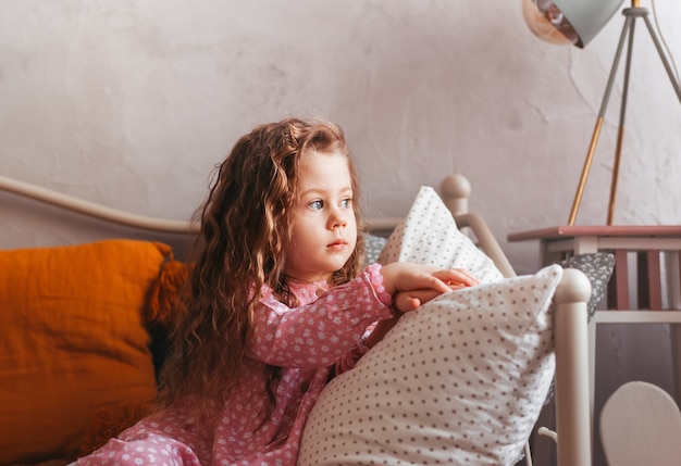 Una niña triste se sienta en una cama en el dormitorio y mira por la ventana. emociones de los niños. Estado de ánimo melancólico