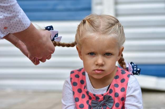 La niña triste se sienta y espera mientras mamá trenza su cabello de su cabello