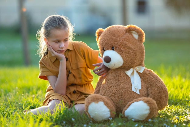 Niña triste y solitaria que pasa tiempo junto con su amigo osito de peluche al aire libre en el soleado patio trasero Soledad en la edad preadolescente