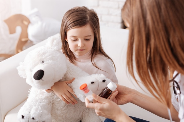 Niña triste sosteniendo su juguete mullido y va a tomar pastillas mientras el médico profesional le da consejos