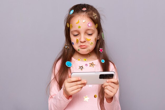 Una niña triste con un teléfono inteligente aislada en un fondo gris cubierta con pegatinas coloridas jugando al juego viendo una lección en línea mirando la pantalla con labios de puchero