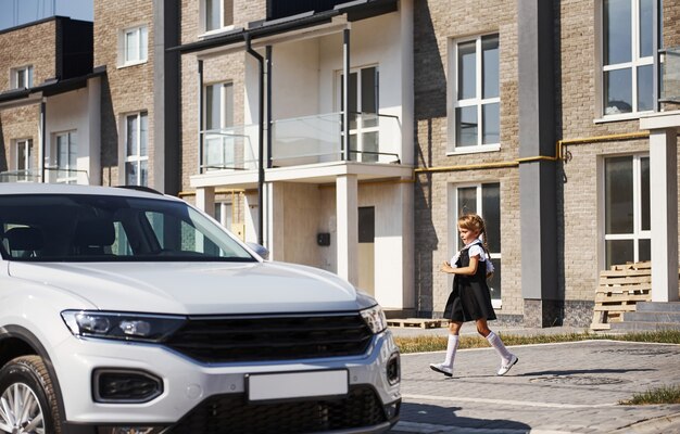 Niña en uniforme escolar corre ro un moderno coche blanco al aire libre en la calle.