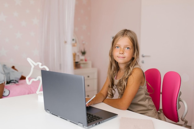 La niña usa un video tutorial en una computadora portátil sentada en una mesa en una habitación y mirando la cámara Capacitación en línea Escuela remota para niños