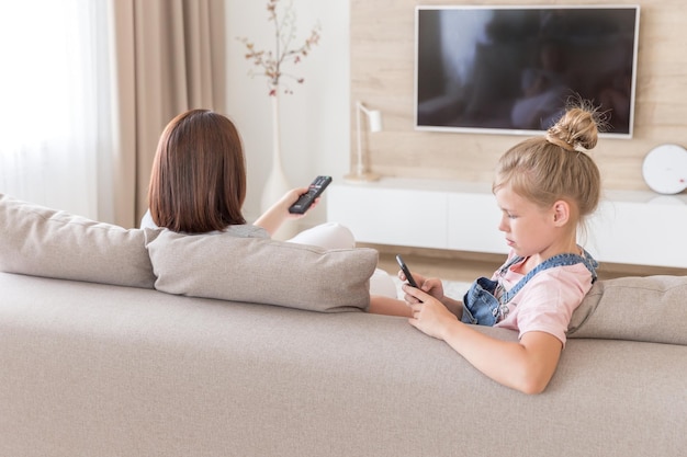 Niña usando teléfono inteligente sentada junto a su madre en el sofá en casa