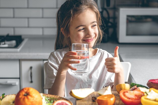 Niña con un vaso de agua en una cocina con frutas y verduras