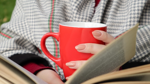 Una niña vestida con un abrigo y una bufanda en el bosque de otoño sostiene un libro y una taza con una bebida caliente en sus manos de cerca en un parque de la ciudad en un día caluroso. El concepto de lectura, relajación y comodidad.