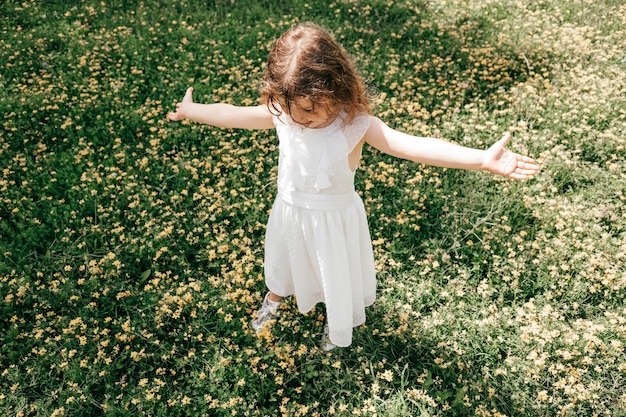 Niña vestida de blanco, con los brazos extendidos, se encuentra en medio de una pradera floreciente. Seguro de vida. Infancia despreocupada. Días de verano. Serenidad. Belleza de la naturaleza. Paseos y aventuras.