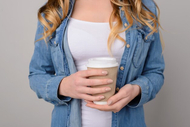 Niña vestida con camiseta blanca y camisa de jeans aislado sobre fondo gris sosteniendo una taza de café para llevar